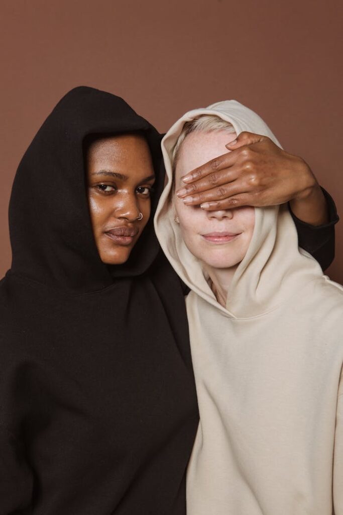 pexels-photo-7479836 African American woman in black hoodie looking at camera while covering eyes of anonymous model on brown background in studio